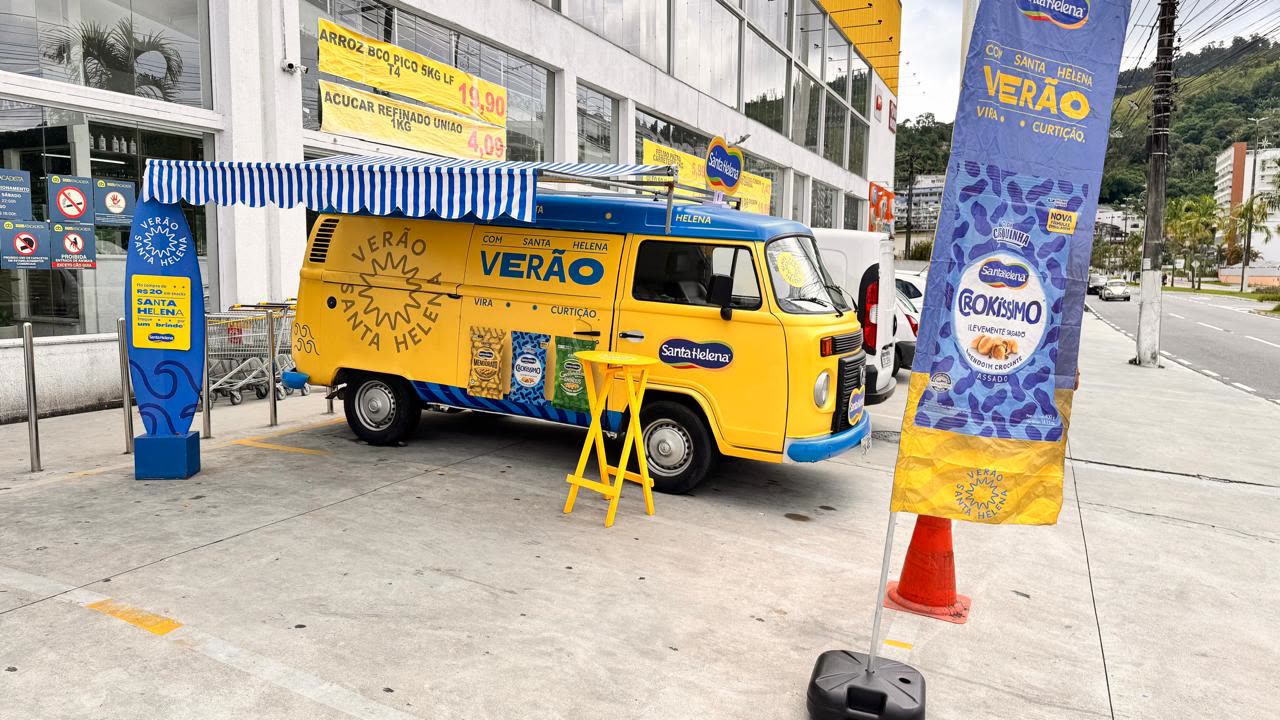 Kombi amarela do Verão Santa Helena com toldo listrado em frente a um supermercado no litoral
